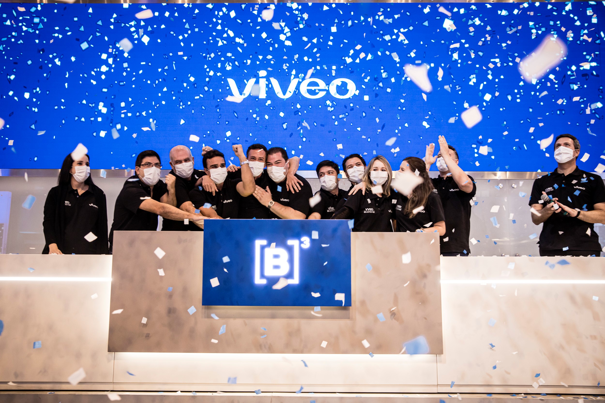 Grupo de pessoas vestindo camisetas pretas com ticker da Viveo unem as mãos em comemoração ao IPO. Ao fundo, logo da Viveo em tela azul com chuva de papel picado.