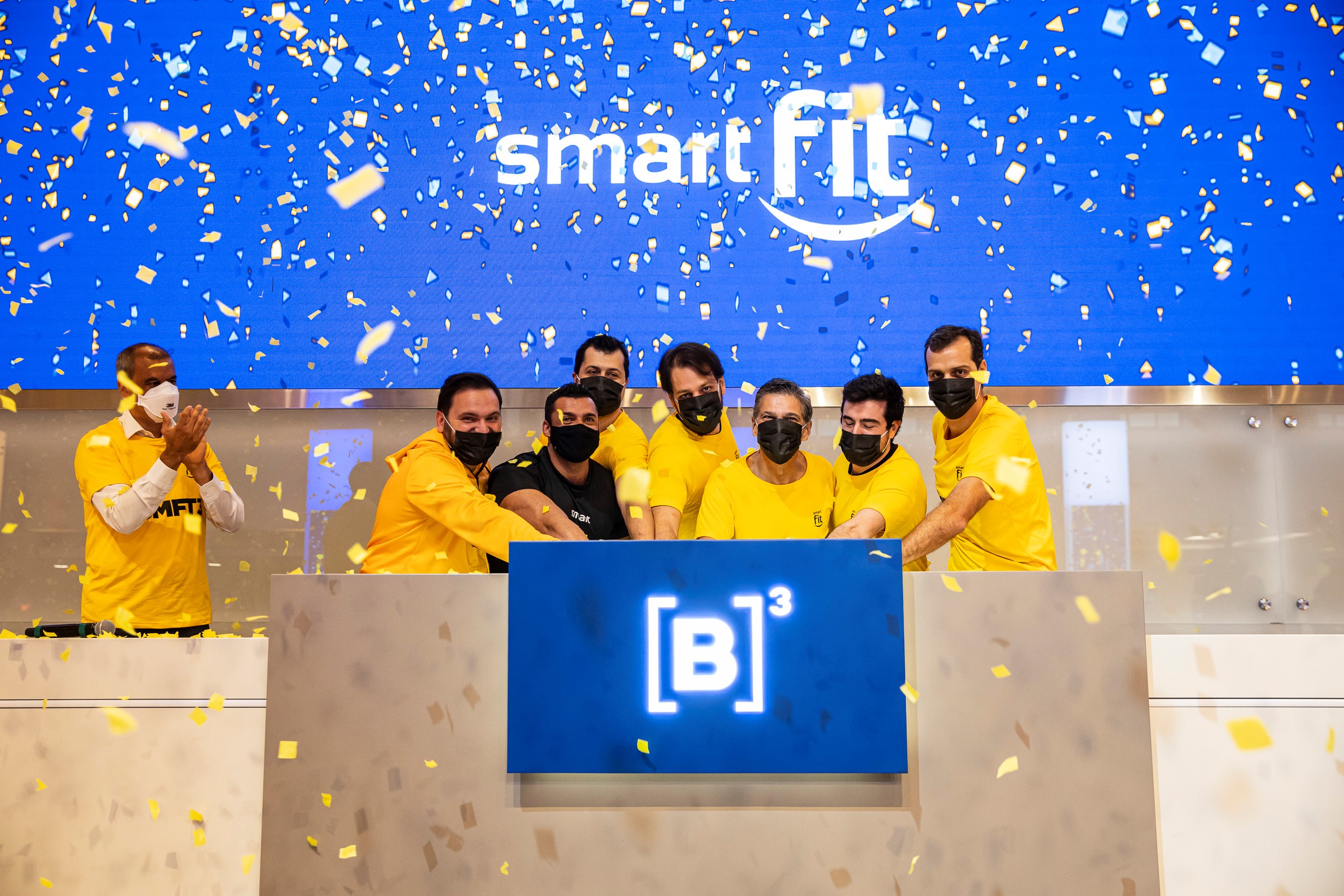 Foto mostra pessoas vestidas com camisetas amarelas e máscaras pretas no momento do toque de campainha. Ao fundo, tela azul com logo da Smart Fit e chuva de papel picado amarelo.