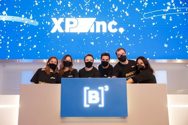 Seis pessoas usando máscara e camista pretas tocam a campainha em comemoração ao início da negociação dos novos ETFs. Ao fundo, tela em azul com confetes brancos e logo da XP Inc.
