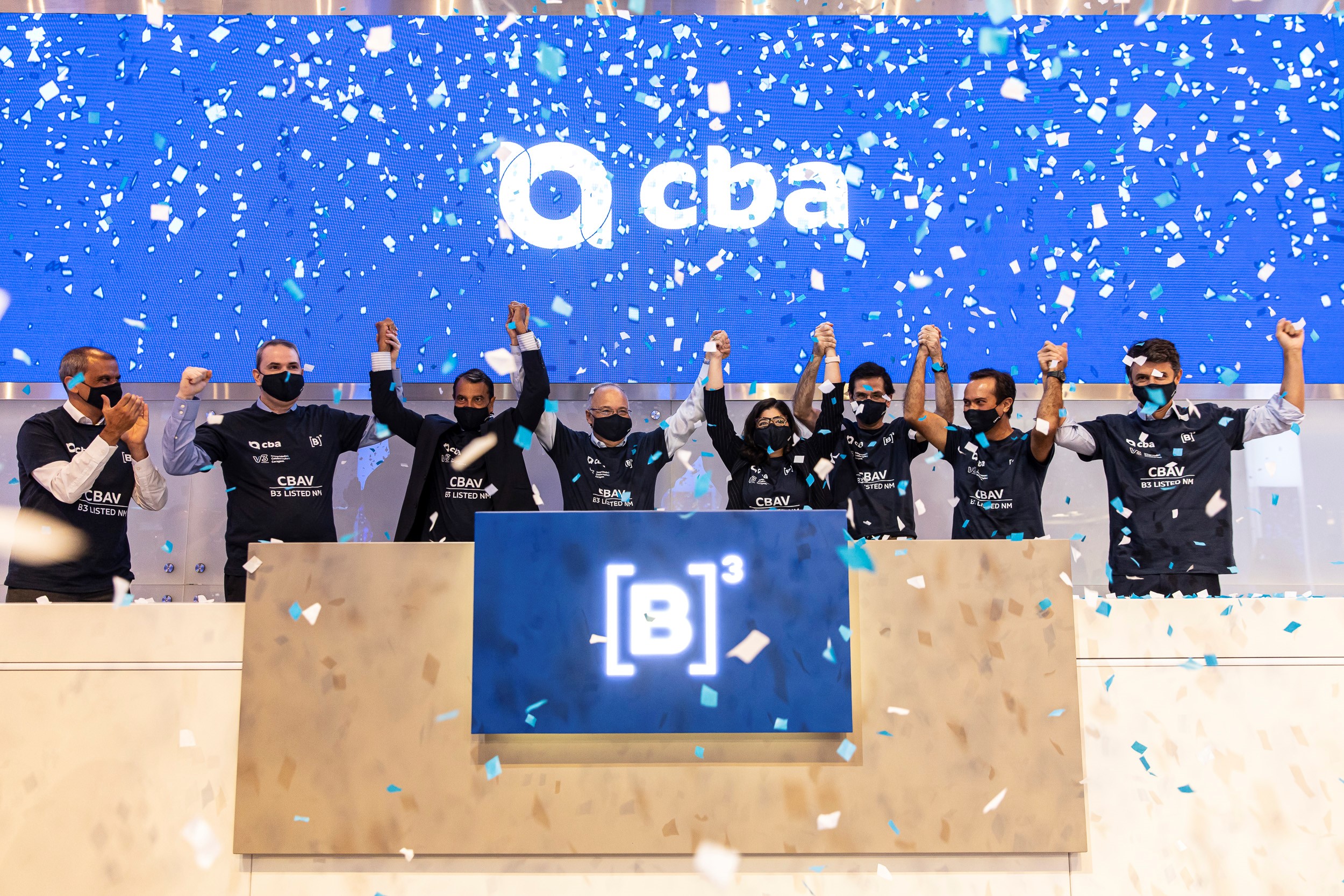 Oito pessoas de camiseta preta com logo da CBA estampado em branco erguem os braços em comemoração ao toque de campainha. Ao fundo, chuva de papel picado prata e um telão azul com logo da CBA.