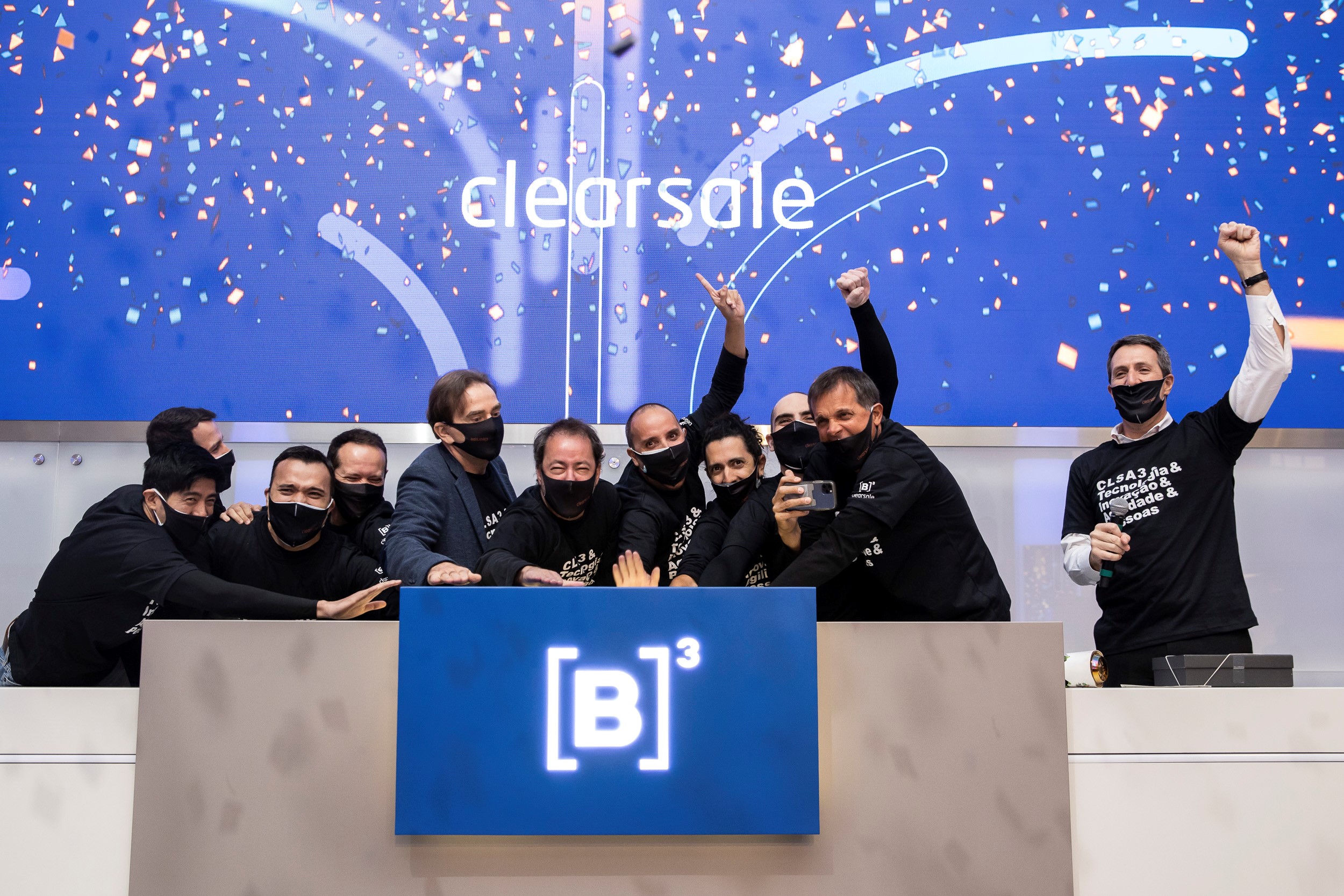 Grupo de homens vestindo camiseta preta com ticker da ClearSale sorriem por baixo das máscaras enquanto tocam campainha. Chuva de papel picado ao fundo marca entrada da ClearSale no mercado de capitais.
