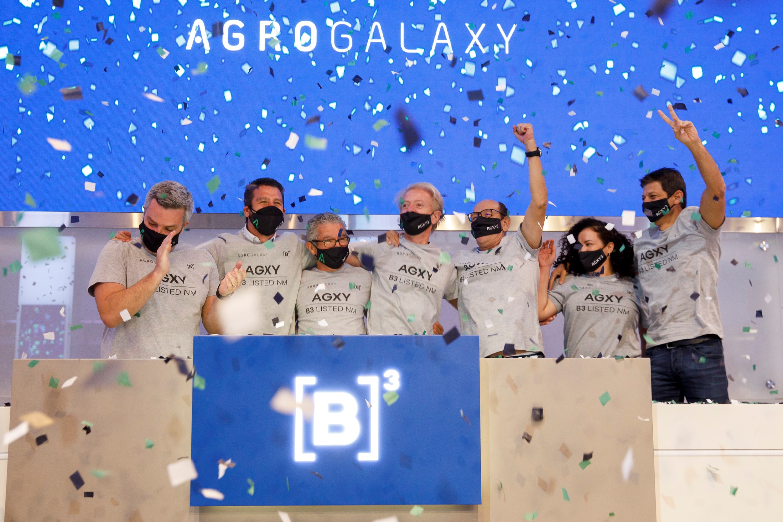 Sete pessoas com os braços estendidos de camiseta cinza com ticker do AgroGalaxy comemoram toque de campainha. Ao fundo, tela azul com chuva de confete e logo do AgroGalaxy.