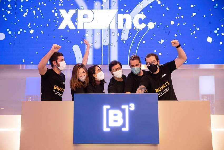 Seis pessoas, todas de máscara, comemoram toque de campainha usando camiseta preta com ticker BOVX11, presente também na tela ao fundo em azul com confetes brancos.
