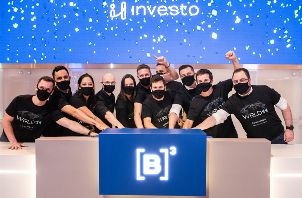 Em cerimônia de toque de campainha, grupo de pessoas une as mãos ao comemorar a estreia do novo ETF. Ao fundo, tela azul com chuva de papel picado e logo da Investo.