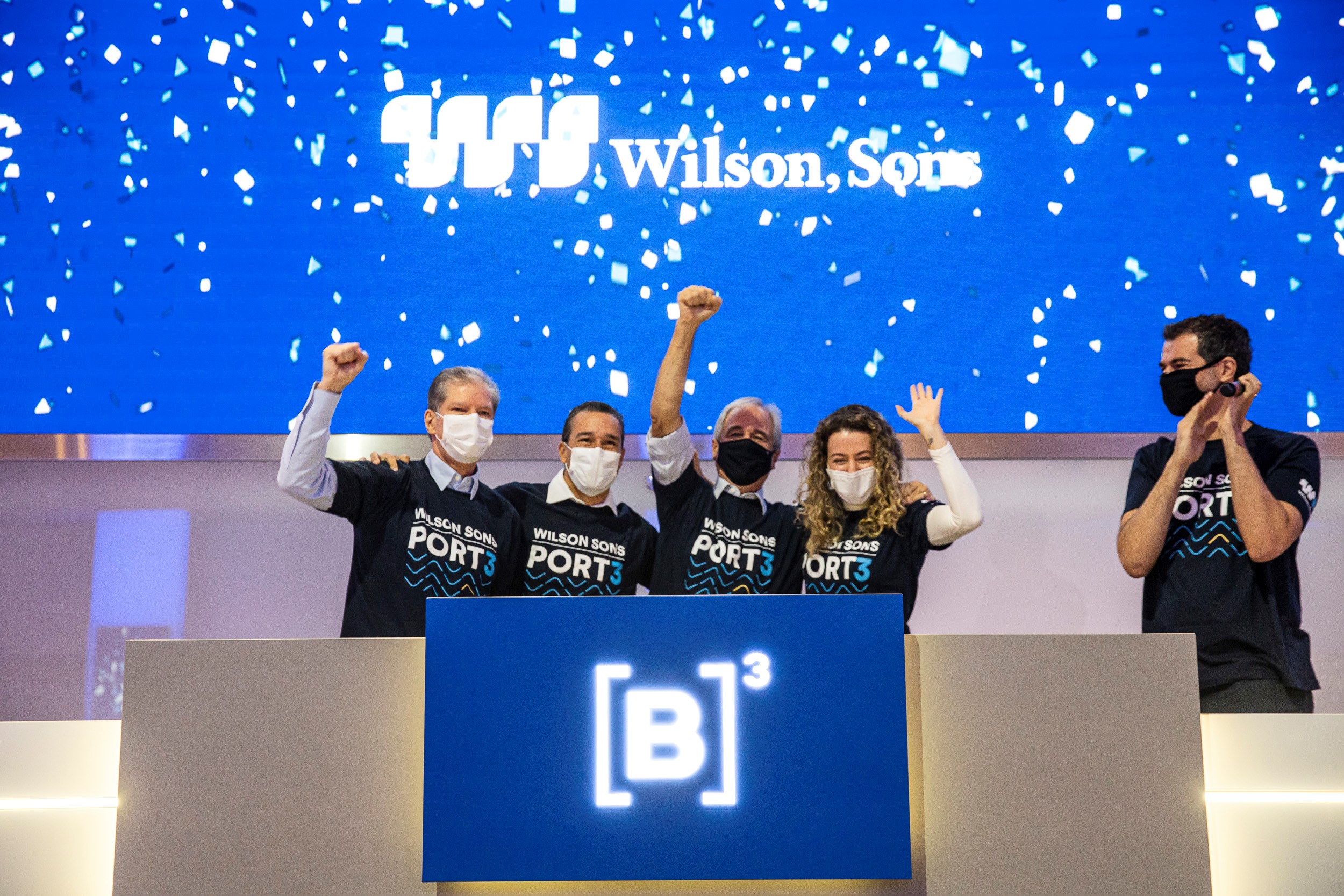 Grupo de pessoas vestindo camisetas pretas com estampa de onda em azul e ticker da wilson sons erguem as mãos em comemoração ao toque de campainha.