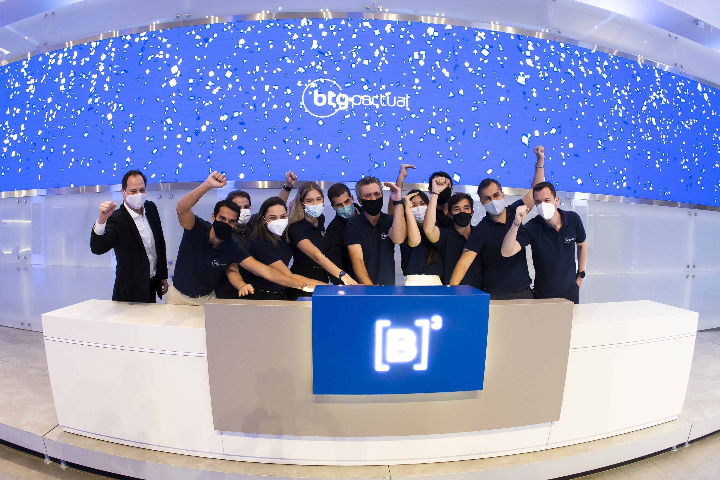 Grupo de pessoas com vestindo camiseta polo preta tocam juntas a campainha. Ao fundo, tela azul com logo da BTG Pactual.
