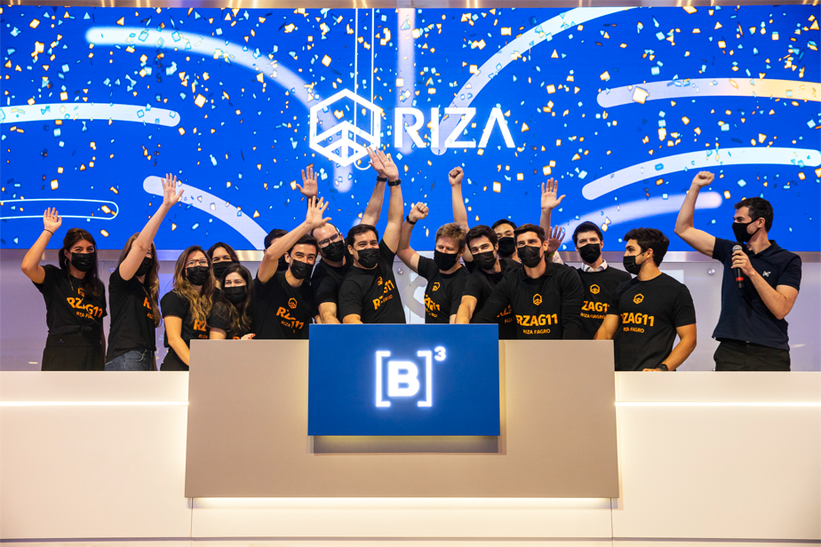 grupo de pessoas ergue as mãos em comemoração ao toque de campainha. Ao fundo, chuva de papel picado dourado em frente a tela azul com logo da Riza.