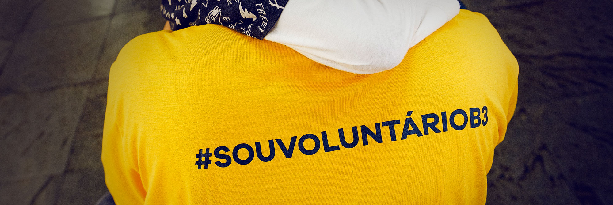 Mostra parte das costas de uma pessoa adulta abaixada, com o braço de uma criança abraçando o seu pescoço. A pessoa adulta veste camisa amarela com o dizer #SOUVOLUNTÁRIOB3