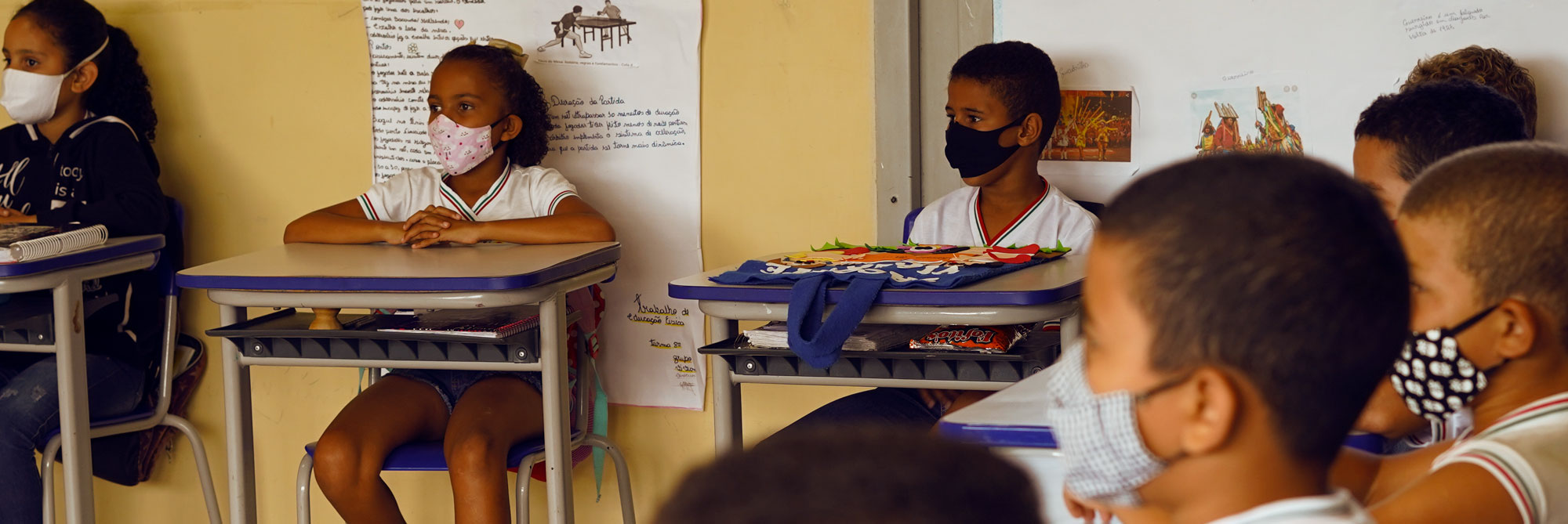 Sala de aula de uma escola pública, sete crianças sentadas, cada uma em sua mesa, olhando para a frente da sala de aula, todas usam máscaras de proteção. A parede da sala de aula é de cor bege e possui cartazes fixados
