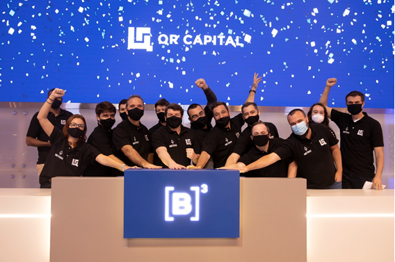 Grupo de pessoas com máscaras e camisetas pretas sorriem em comemoram juntas toque de campainha. Ao fundo, tela azul com logo da QR Capital e chuva de papel picado.