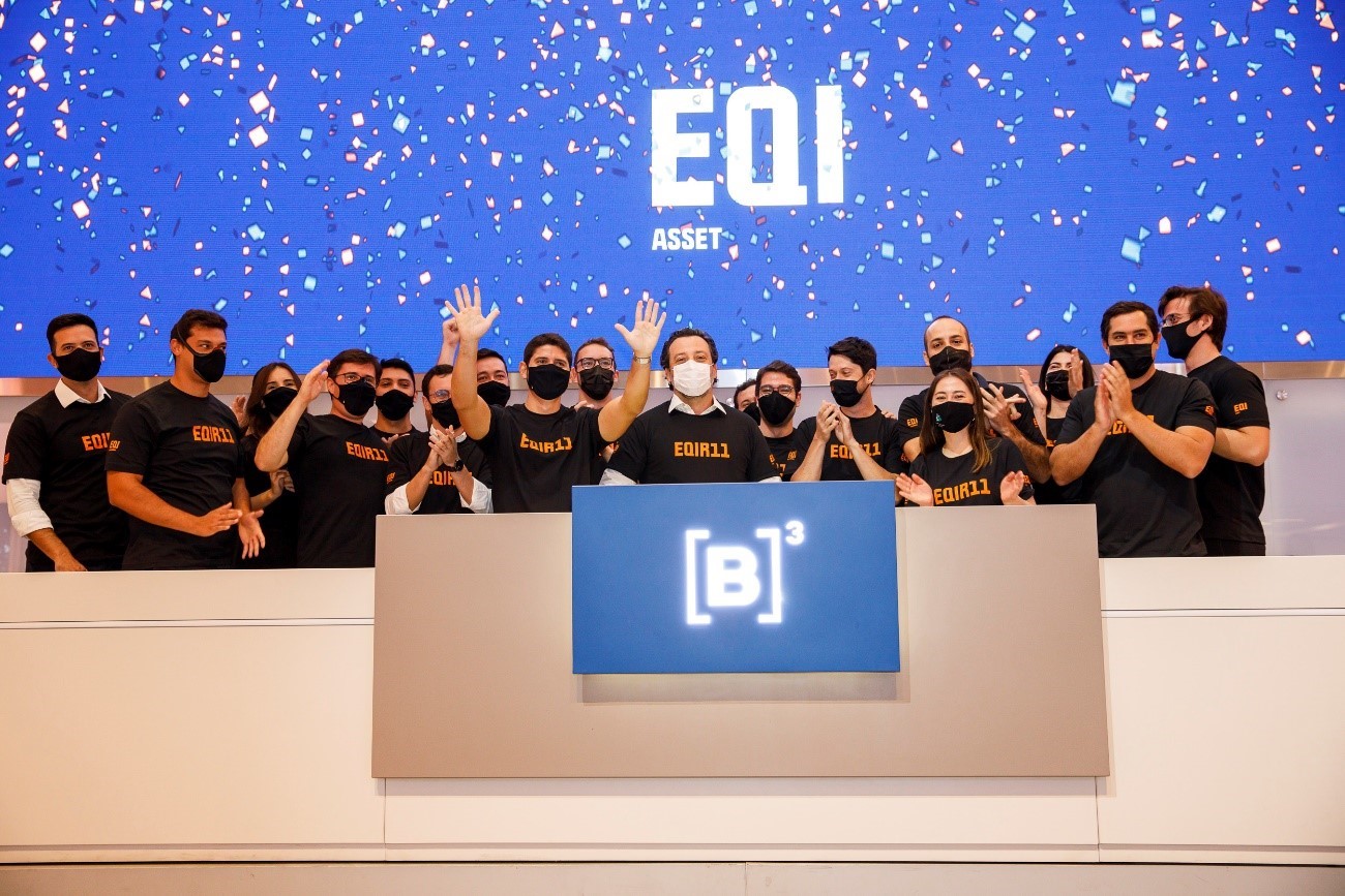 Grupo de pessoas atrás do púlpito da B3 se unem em comemoração ao toque de campainha que marca o início da negociação das cotas do fundo. Na tela ao fundo, chuva de papel picado e logo da EQI