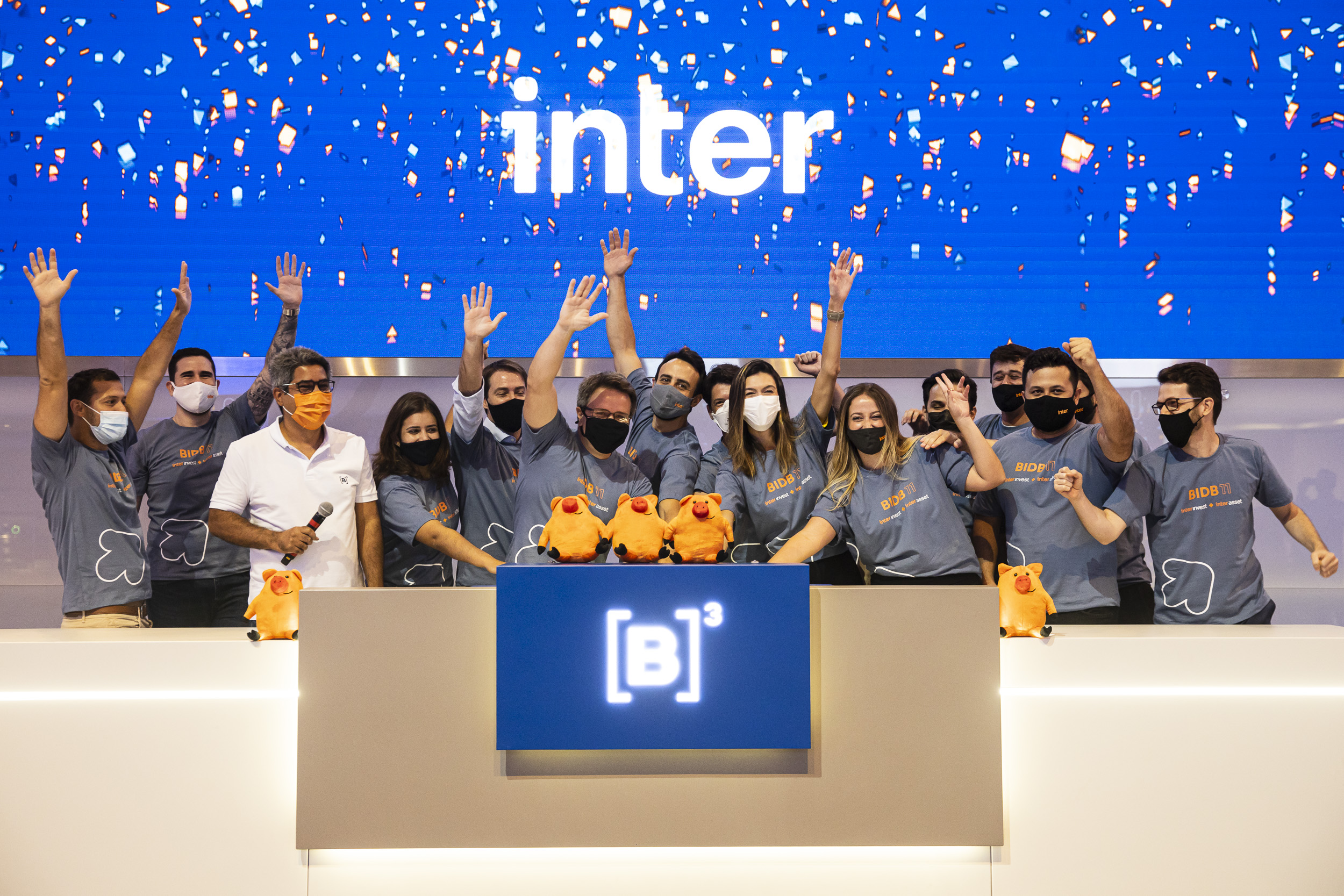 Grupo de pessoas ergue as mãos comemorando toque de campainha. Ao fundo, chuva de papel picado laranja e azul em frente a tela azul com logo do Inter.