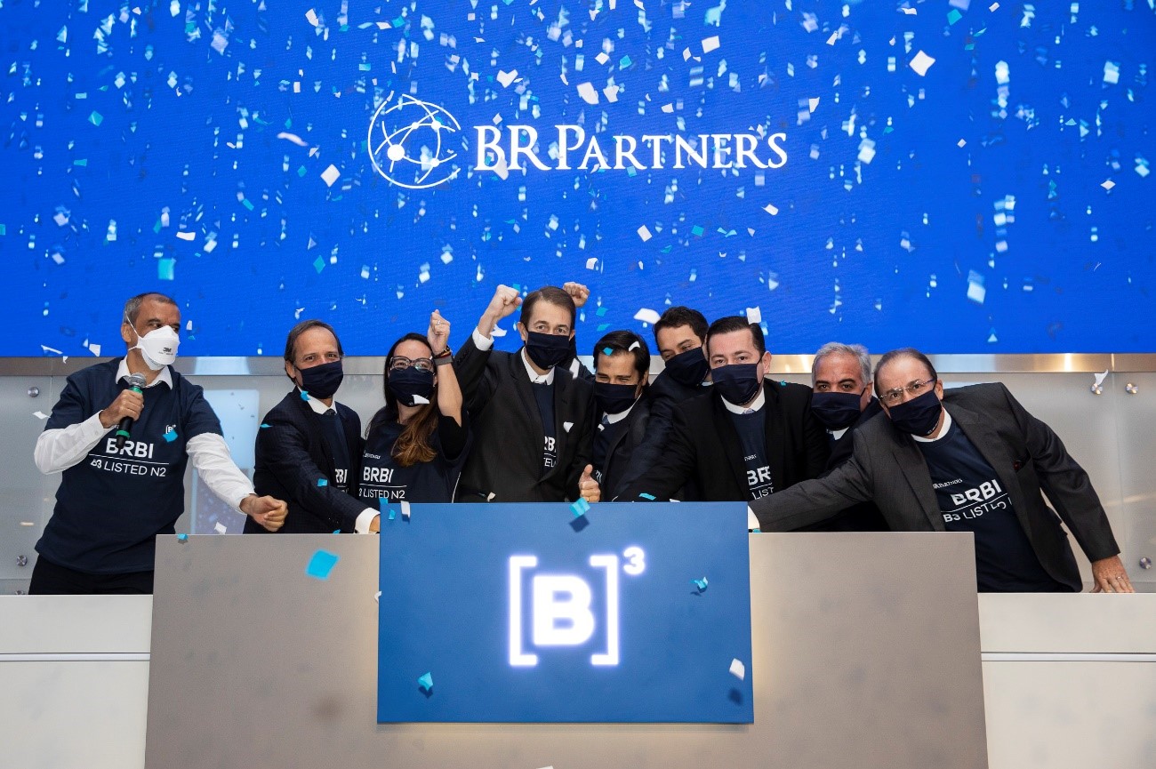 Nove pessoas reunidas de máscara atrás do púlpito tocam a campainha comemorando o IPO da BR Partners. Ao fundo, telão em azul com confetes brancos e logo da BR Partners.