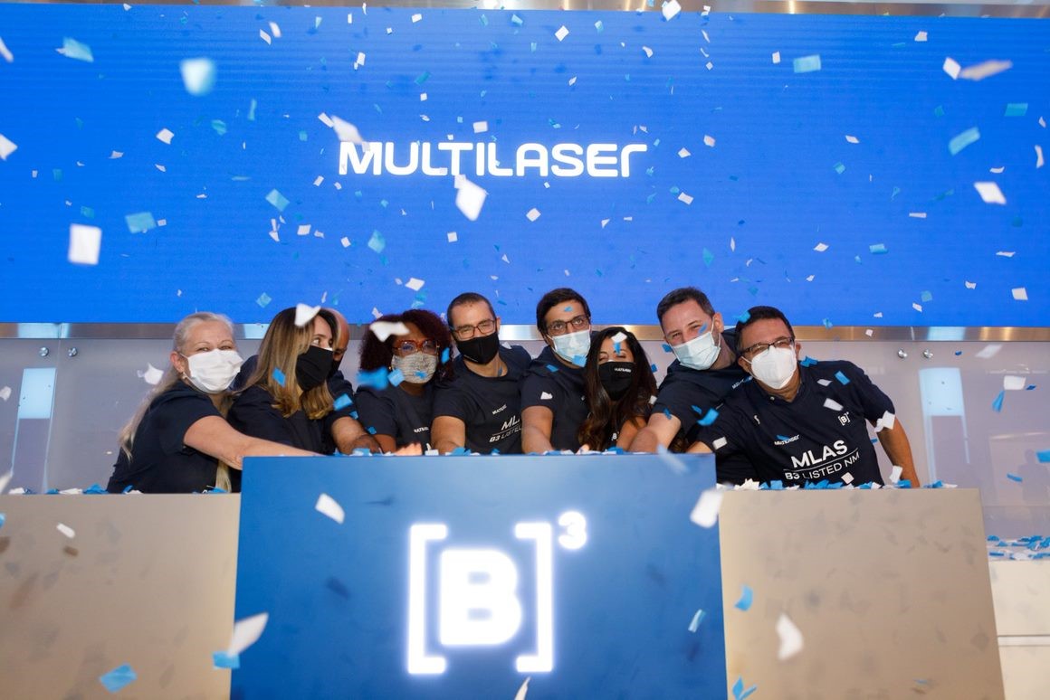 Nove pessoas com camisetas pretas com logo da Multilaser e de máscara tocam a campainha comemorando IPO.Ao fundo, chuva de papel picado e tela azul com logo da Multilaser.