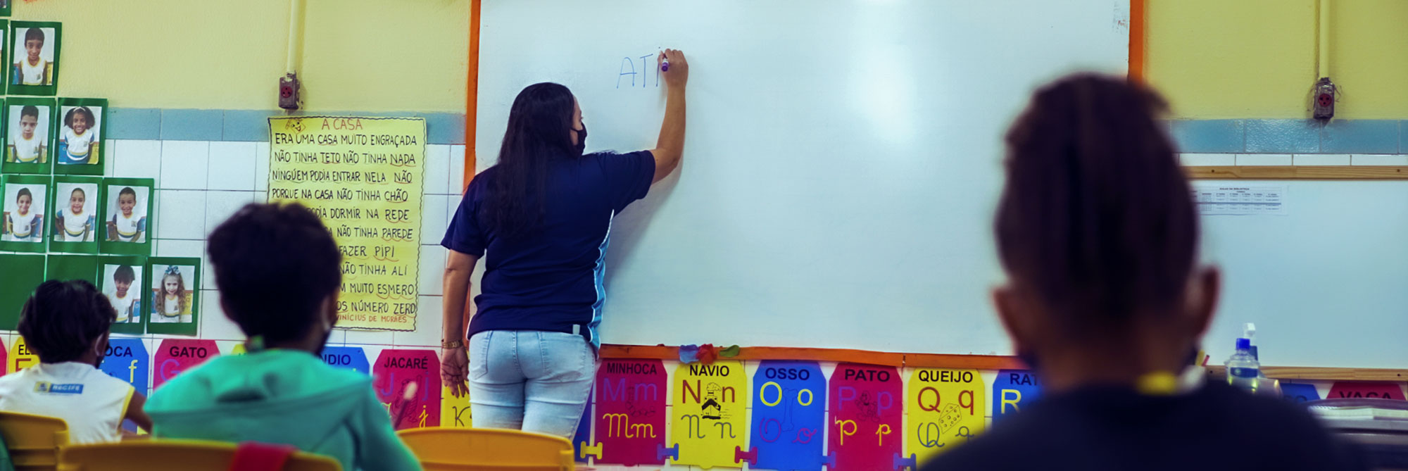 Sala de aula de uma escola pública, com muitos cartazes colados na parede. Uma professora, mulher adulta com altura mediana cabelos pretos médios, usando camiseta azul e calça jeans azul claro, aparece de costas escrevendo no quadro branco. À esquerda, na parede, existe um painel verde com fotos de crianças. Observando a professora estão três crianças de costas, sentadas: uma mais à esquerda com camiseta branca, ao seu lado direito uma segunda criança com blusa de frio verde clara, com cabelos pretos, à direita da foto a terceira criança, de pele negra e cabelos pretos, vestindo blusa azul escura