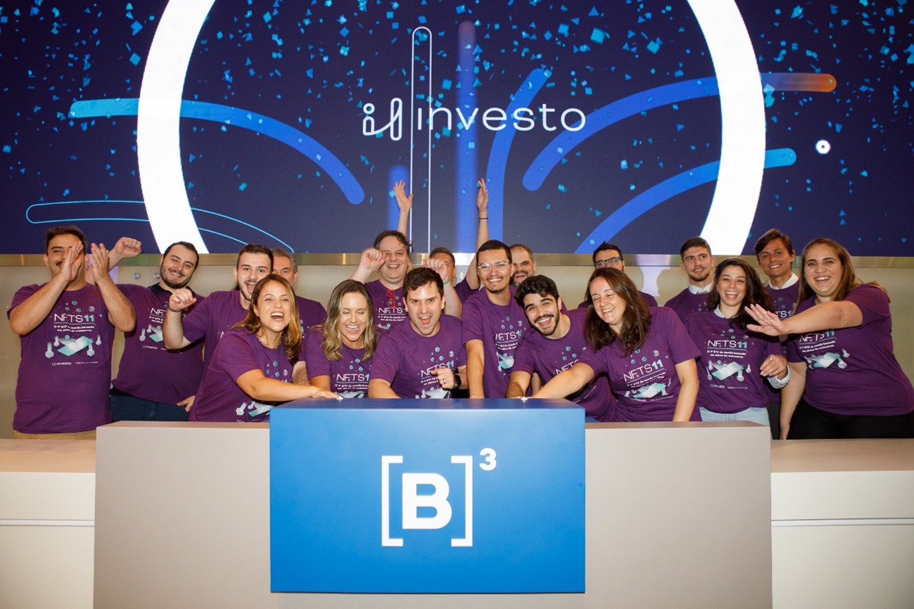 Grupo de pessoas se reúne no púlpito para comemorar lançamento do produto. Elas erguem as mãos e batem palmas em frente ao telão com logo da investo.