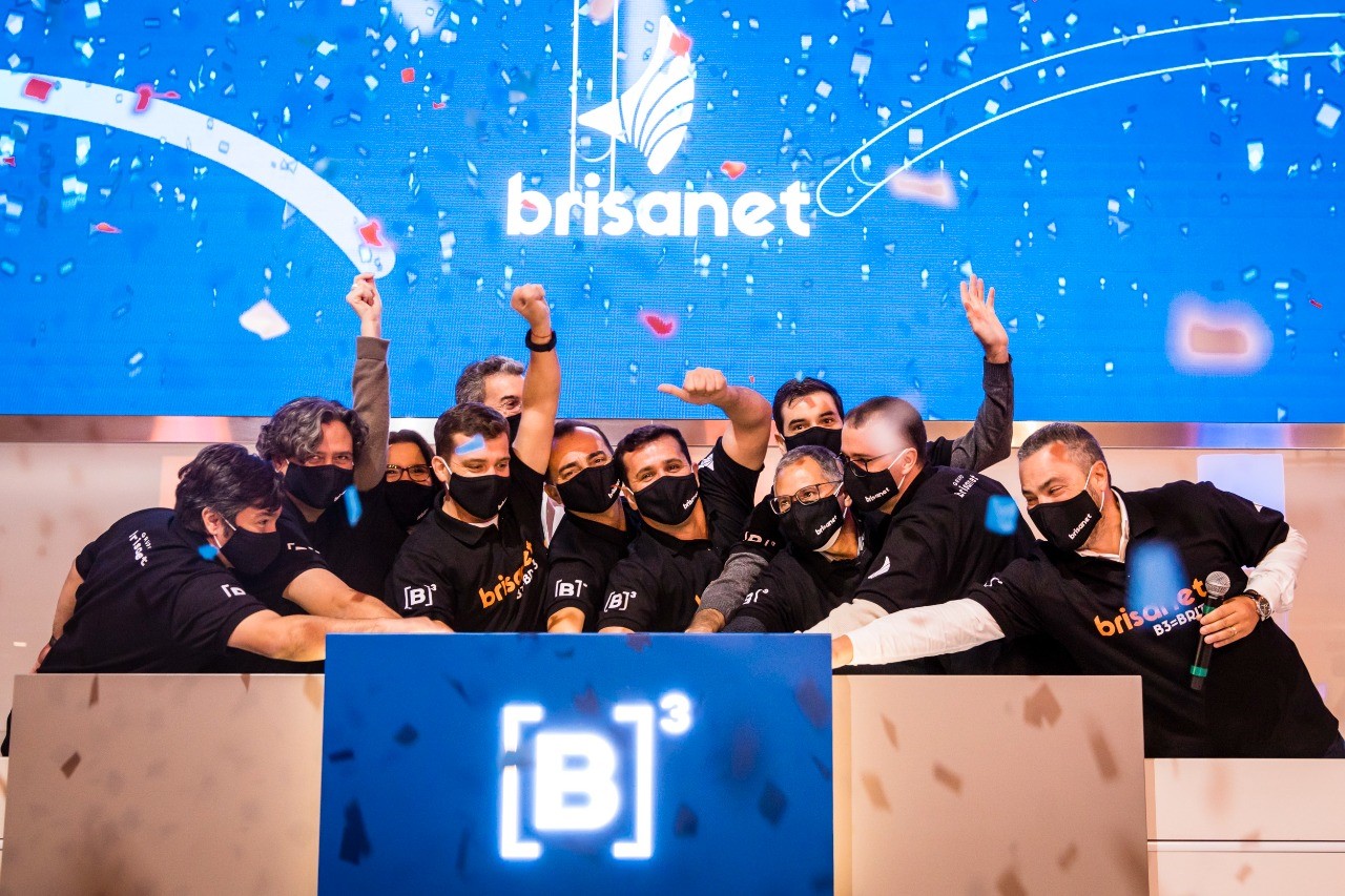 Grupo de pessoas com camiseta preta com ticker e nome da Brisanet se unem em toque de campainha. Ao fundo, chuva de papel picado em tela azul com logo da empresa.