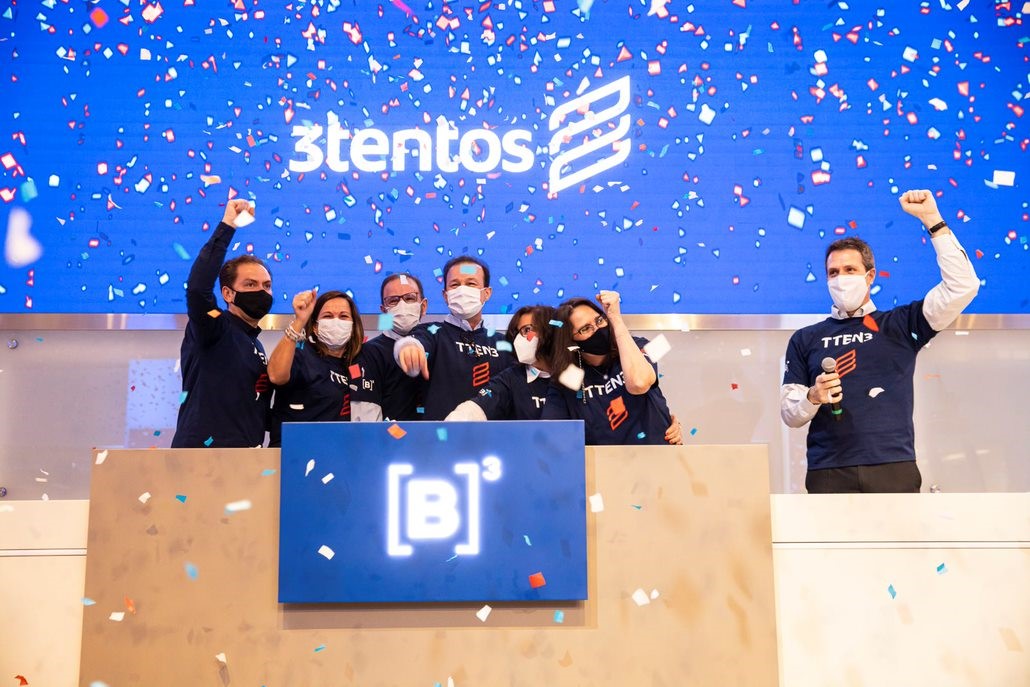 Sete pessoas, vestindo camiseta azul marinho com logo da 3Tentos, levantam as mãos em comemoração ao toque de campainha. Ao fundo, telão em azul com chuva de papel picado e logo da 3Tentos.