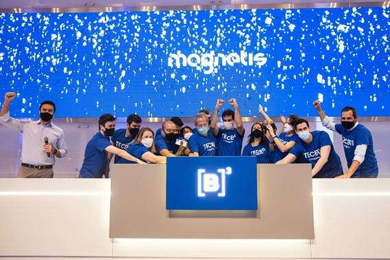 Grupo de pessoas vestindo camisetas azuis se unem em toque de campainha comemorativo. Ao fundo, tela azul com chuva de papel picado branco e logo da Magnetis.