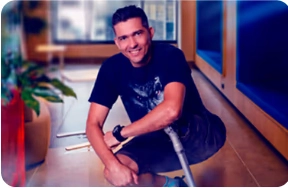 Smiling man kneeling on the floor with a prosthetic right leg in a bright indoor setting.