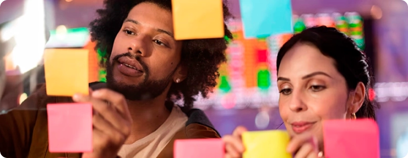 Two people are placing colorful Post-it notes on a glass wall. The man on the left has curly hair and a beard, and the woman on the right has long dark hair tied back. They appear focused and collaborative, with blurred background lights suggesting an organized environment.
