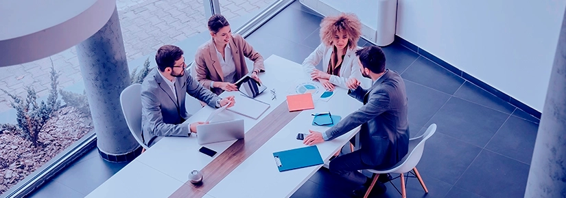 Four people in a business meeting around a table, with laptops and documents. The environment is modern and well lit.