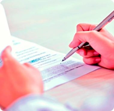 Close-up of a person holding a pen and writing on a document. The document is slightly out of focus and another hand is visible holding the paper firmly. The background is a soft, blurred gradient of pink and white tones.